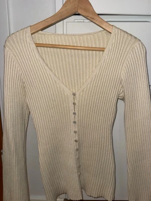 Beige ribbad kofta med knappar - Snygg beige ribbstickad kofta med v-ringning och långa ärmar. Koftan har flera matchande knappar framtill och är figurnära i modellen. Perfekt att styla med jeans eller kjol för en clean look.