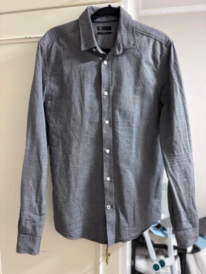 Grå skjorta - Säljer en stilren grå skjorta från Cubus i regular fit. Skjortan har klassisk krage, vita knappar framtill och långa ärmar. Perfekt för dig som gillar en enkel och clean look. Längd:68 cm, bredd: 47 cm