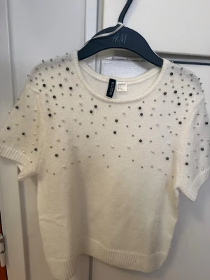 Vit pärlprydd kortärmad stickad tröja  - Kortärmad vit stickad topp från H&M, dekorerad med små pärlor i silver och vitt över axlar och bröst. Rund halsringning och ribbade kanter ger en snygg och trendig look. Perfekt för dig som gillar detaljer!