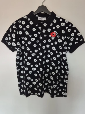 Svart blommig piké från WESC - Svart piké från WESC med klassiskt krage och korta ärmar. Piké har ett vitt blommigt mönster över hela plagget och en röd detalj på bröstet. Tillverkad i mjuk bomull som känns skön mot huden. Perfekt för dig som gillar statement-plagg.