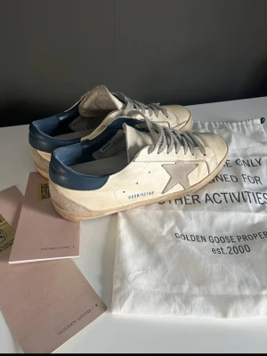 Golden Goose Superstar sneakers beige/blå - Golden Goose Superstar sneakers i beige mocka och läder med blå häl och grå stjärndetalj på sidan. Klassisk låg modell med slitna detaljer och vita snören. Snygg vintagekänsla och ikonisk design som sticker ut.