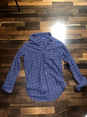 Blårutig vintage skjorta från Ralph Lauren - Klassisk blårutig vintage skjorta från Ralph Lauren med knappar framtill och lång ärm. Skjortan har en liten röd logga broderad på bröstet och är tillverkad i mjuk bomull. Perfekt för en avslappnad men ändå stilren look.