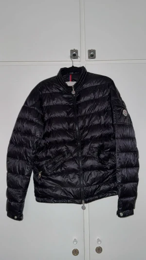 Svart Moncler dunjacka herr - Säljer en svart Moncler dunjacka med klassisk quiltad design och rund krage. Jackan har dragkedja framtill, fickor med dragkedja och Moncler-logga på ärmen. Perfekt för kyliga dagar och har en clean, stilren look. 