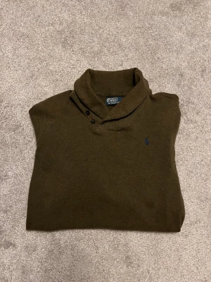 Olivgrön tröja från Polo Ralph Lauren - Snygg olivgrön tröja från Polo Ralph Lauren med lång ärm och klassisk krage med knappar. Tröjan har en diskret broderad logga på bröstet och är tillverkad i mjukt material som passar perfekt till höst och vinter.
