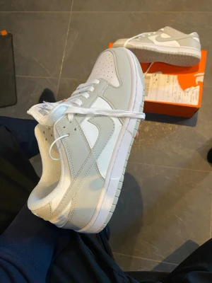 Nike Dunk Low vita sneakers - Säljer ett par fräscha Nike Dunk Low sneakers i vitt och ljusgrått. Skorna har klassisk låg siluett, perforerad tåbox och vit sula. Tillverkade i läder med textildetaljer och ikoniska swoosh-loggan på sidan. Perfekta för dig som gillar stilrena och tidlösa sneakers.