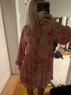 Klänning - Söt kortklänning i rosa och vitt med blommigt mönster och v-ringning. Klänningen har långa ärmar, volangdetaljer och knyt i midjan. Perfekt för varmare dagar och har en luftig passform.