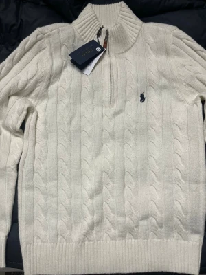 Vit kabelstickad tröja Polo Ralph Lauren - Kabelstickad vit tröja från Polo Ralph Lauren med halv dragkedja och hög krage. Tröjan har det klassiska broderade logotypen på bröstet och ribbstickade muddar. Perfekt för lager-på-lager och snygg till jeans eller chinos.