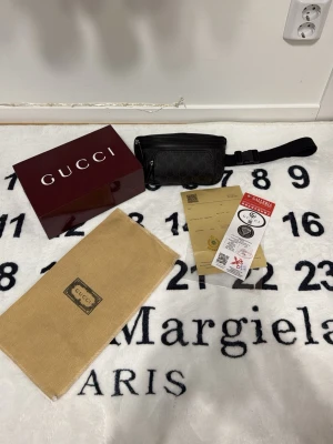 Svart axelväska från Gucci - Snygg svart axelväska från Gucci med klassiskt GG-mönster i canvas och detaljer i läder. Väskan har justerbar rem, dragkedja framtill och medföljer originalbox, dustbag och äkthetsintyg. Perfekt för dig som vill ha en stilren och exklusiv accessoar.