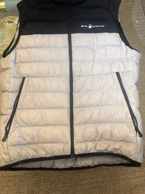 Grå och svart dunväst Sail Racing - Snygg dunväst från Sail Racing i grått och svart. Västen har två dragkedjeförsedda fickor framtill, hög krage och quiltad design. Perfekt för lager-på-lager och håller dig varm med lättviktigt dunmaterial. Logga på bröstet för en sportig look.