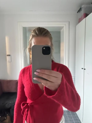 Röd omlottkofta med knytband - Snygg röd omlottkofta med knytband i midjan och långa ärmar. Koftan har en mjuk och fluffig känsla, perfekt för att ge en trendig look. Passar till både jeans och kjol för en cool stil.