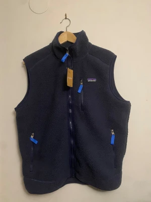 Mörkblå fleeceväst från Patagonia - Mysig mörkblå fleeceväst från Patagonia med hög krage och tre fickor med blå dragkedjor. Västen har en klassisk passform och är perfekt för lager-på-lager. Patagonia-logga på bröstet och mjukt material som håller dig varm.
