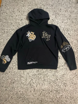 Svart hoodie  från Supply - Svart hoodie från Supply med stora tryck i guld och vitt på bröstet, ärmarna och fickan. Motiven inkluderar ett S med en ängel, NY-text och flera coola detaljer. Hoodien har huva och en stor magficka framtill. Perfekt för dig som gillar streetwear och unika prints.