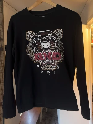 Svart Kenzo hoodie med tigertryck - Säljer en svart hoodie från Kenzo med det ikoniska tigerhuvudet broderat framtill och texten 'KENZO PARIS' i vitt och rött. Tröjan har rund halsringning och långa ärmar. Perfekt för dig som vill sticka ut med en streetwear-look.