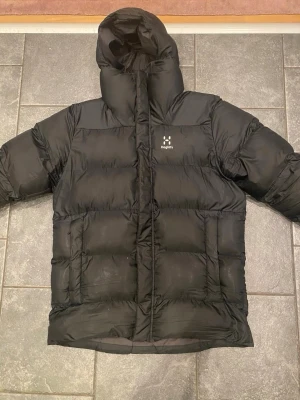 Svart pufferjacka från Haglöfs - Snygg svart pufferjacka från Haglöfs med stor huva och quiltad design. Jackan har en hög krage och diskret Haglöfs-logga på bröstet. Perfekt för kalla vinterdagar och riktigt varm tack vare det tjocka materialet. Klassisk och stilren look.