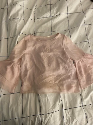 Ljusrosa blus med volangärmar - Söt ljusrosa blus i transparent, lätt material med vida volangärmar och rund halsringning. Blusen har en lös passform och är perfekt för en trendig och feminin look. Storleken är egentligen 146 men passar som xxs/xs enligt mig.