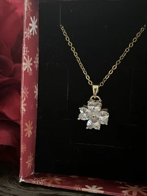 Guldigthalsband  med blomformad kristall - Snyggt halsband med tunn guldfärgad kedja och ett hängsmycke i form av en blomma med gnistrande, klara kristaller. Perfekt för dig som gillar eleganta och stilrena accessoarer med lite extra bling.