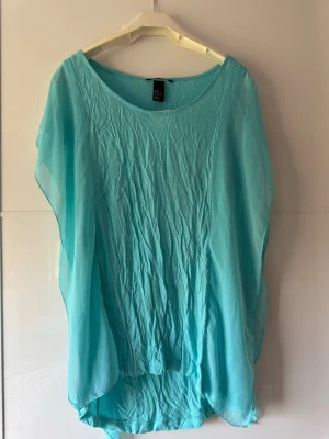 Ny.Turkos blus med fladdriga ärmar - Säljer en turkos blus från H&M med lös passform och fladdriga, transparenta ärmar. Toppen har rund halsringning och är tillverkad i ett tunt, lätt material som ger en luftig känsla. Perfekt för varma dagar och enkel att styla.