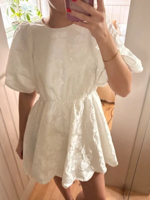 Vit kort klänning med puffärmar - Superfin vit kort klänning med puffiga ärmar och ett vackert blommigt mönster. Klänningen har en öppen rygg med scallop-detaljer och markerad midja som ger en snygg siluett. Perfekt för dig som vill sticka ut med en unik och trendig look.