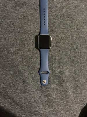 Apple Watch med blått silikonband - Smartklocka från Apple med rektangulär silverfärgad aluminiumram och svart skärm. Klockan har ett stilrent blått silikonband med justerbara hål och metallspänne. Perfekt för dig som vill ha koll på träning, notiser och hälsa direkt på handleden.