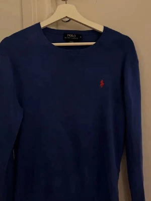 Blå tröja från Polo Ralph Lauren - Snygg blå långärmad tröja från Polo Ralph Lauren i slim fit-modell. Tillverkad i mjuk pima cotton och har den klassiska röda loggan broderad på bröstet. Perfekt för en stilren och avslappnad look.