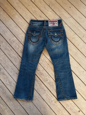 True Religion Y2K bootcut distressed washed jeans - Ett par True Religion jeans med en klassisk design. Jeansen har detaljerade sömmar i olika färger, inklusive orange och blått, som framhäver fickorna och sidorna.   Midjan har bälteshällor och en knappgylf. Jeansen har en lätt sliten look som ger dem karaktär.  Knappen saknas på vänster bakficka. Putte litet hål i sidan på höger byxben. Annars i fint skick.  Mått: Midja 39 cm, längd 92 cm