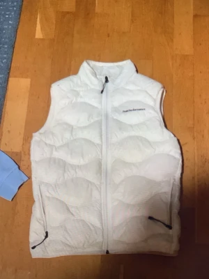 Vit dunväst från Peak Performance - Snygg vit dunväst från Peak Performance med hög krage och dragkedja framtill. Västen har två fickor med dragkedja och diskret logga på bröstet. Perfekt att slänga över en hoodie eller tröja för extra värme. Klassisk och stilren design.