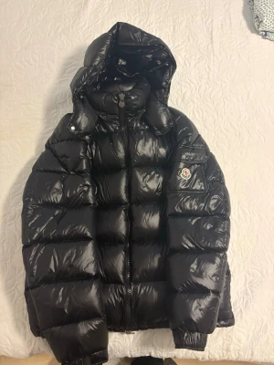 Moncler Maya - Säljer en svart, glansig dunjacka från Moncler med avtagbar huva och klassisk logotyp på ärmen. Jackan har breda kanaler, dragkedja framtill och ribbade muddar vid ärmslut. Perfekt för kalla vinterdagar och riktigt snygg streetstil.