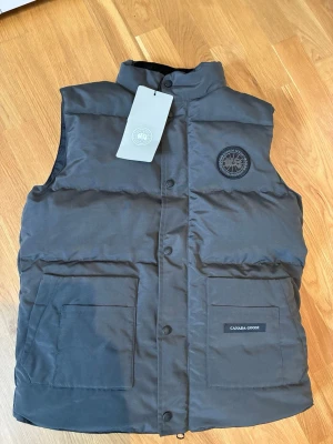 Canada Goose väst Helt Ny!!! - Snygg svart dunväst från Canada Goose med broderad logotyp på bröstet och två stora fickor framtill. Västen har dragkedja och tryckknappar samt hög krage. Perfekt för lager-på-lager och håller dig varm under kyliga dagar.