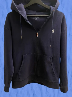 Polo Ralph Lauren zip hoodie  - Mörkblå hoodie med dragkedja från Polo Ralph Lauren. Klassisk design med huva och dragsnören, två fickor framtill och den ikoniska vita polospelaren broderad på bröstet. Tillverkad i mjuk bomullsblandning som känns skön mot huden. Pris kan diskuteras vid snabb affär!!!