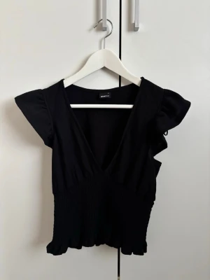 Svart v-ringad topp med volangärm - Snygg svart topp från Gina Tricot med v-ringning och volangärmar. Modellen är figurnära med smock i midjan och lätt rynkad nederkant. Perfekt att styla med jeans eller kjol för en trendig look.