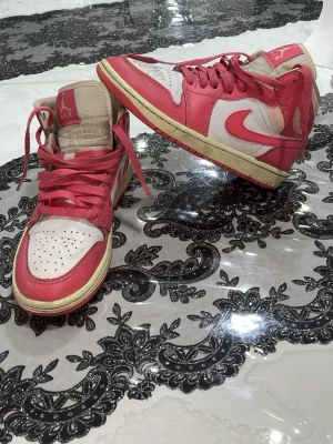 Air Jordan 1 Mid rosa/vit - Säljer ett par Air Jordan 1 Mid sneakers i rosa och vitt läder med beige detaljer. Klassisk Jordan-logga på sidan och perforerad tåbox. Skorna har rosa snören och en retro vibe som sticker ut.