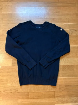 Mörkblå stickad tröja från Pelle P - Mörkblå stickad tröja från Pelle P med rund halsringning och ribbade muddar vid ärmslut och nederkant. Tröjan har en diskret logodetalj på axeln och är tillverkad i mjukt material som passar perfekt till vardagslooken.