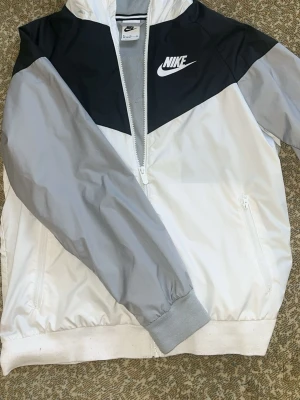 Nike vindjacka i vitt och grått XL Barn - Nike vindjacka med dragkedja i vitt, ljusgrått och svart. Jackan har två sidofickor med dragkedja, ribbade muddar och meshfoder på insidan. Stor Nike-logga på bröstet och huva. Perfekt för en sportig och avslappnad stil.