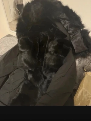 Svart pälsjacka från Roco Stockholm - Säljer en svart pälsjacka från Roco Stockholm med fluffig fuskpäls och stor huva. Jackan har en lyxig look med mjukt material och breda slag. Perfekt för dig som vill ha en statementjacka till vintern.