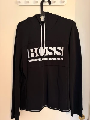 Svart Hoodie Hugo Boss - Svart hoodie från Hugo Boss med stor vit logga på bröstet och vita dragsnören. Storlek L, säljer på grund av att den inte kommer till användning längre. Skriv för mer frågor!