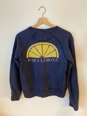 Sweatshirt Acqua Limone XXS - Mörkblå sweatshirt från Acqua Limone med klassisk logga framtill och stort citrongult tryck på ryggen. Tröjan har rund hals, ribbade muddar och är tillverkad i mjukt bomullsmaterial. Perfekt för en avslappnad och trendig stil. Märkt med storlek XXS men passar även en XS. 