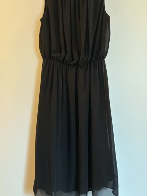 Svart ärmlös klänning i chiffong - Säljer en svart, ärmlös klänning med resår i midjan och lätt, flytande chiffongmaterial. Klänningen har en enkel och stilren design med mjukt fall och rundad halsringning. Perfekt för dig som gillar minimalistisk stil.