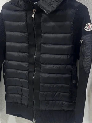 Svart pufferjacka från Moncler, cardigan  - Snygg svart pufferjacka från Moncler med quiltad design och ribbade partier på ärmar och sidor. Jackan har dragkedja framtill, hög krage och Moncler-logga på ärmen. Materialet är en mix av blankt nylon och stickad textil för extra komfort.