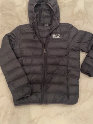 Svart dunjacka från Emporio Armani EA7 - Snygg svart dunjacka från Emporio Armani EA7 med huva och dragkedja framtill. Jackan har ett stilrent EA7-tryck på bröstet. Det är ett litet litet hål uppe vid fickan som inte påverkar fickan på något sätt. Hör av er om ni har någon fråga och skicka gärna bud till mig om inte priset inte stämmer