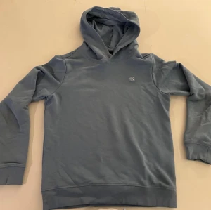 Blå hoodie med huva från Calvin Klein  - Snygg blå hoodie från calvin Klein med klassisk huva och ribbade muddar vid ärmslut och nederkant. Hoodien har en enkel design med en liten logotyp på bröstet och är gjord i mjukt bomullsmaterial. Perfekt för en avslappnad och stilren look.