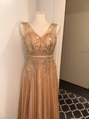 Champagnefärgad långklänning med paljetter - Elegant långklänning i champagnefärg med v-ringning och glittriga paljettdetaljer över hela livet och kjolen. Klänningen har ärmlös design och markerad midja, perfekt för speciella tillfällen. Materialet är lätt och ger ett lyxigt intryck.