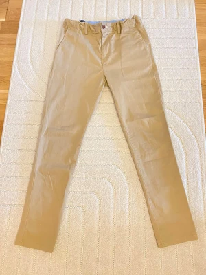 Beige chinos från Gant - Snygga beige chinos från Gant med klassisk rak passform och diskreta fickor både fram och bak. Tillverkade i mjukt bomullstyg med knappstängning och bälteshällor. Perfekta för en clean och stilren look. Köpte den för skolfoto sen dess har jag aldrig haft på mig den har bara hängt den i garderoben 