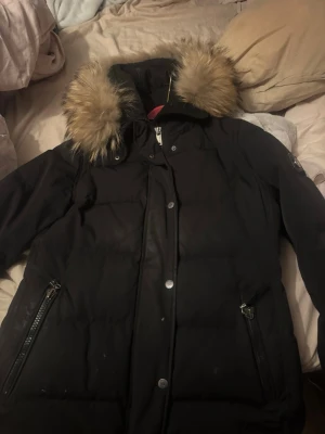 Svart dunjacka med pälskrage från Hollies - Snygg svart dunjacka från Hollies med stor huva och fluffig pälskant. Storlek 40. Jackan har två dragkedjeförsedda fickor framtill och en patch med Hollies-logga på ärmen. Perfekt för kalla vinterdagar och riktigt varm tack vare dunfyllningen. Äkta Päls och väldigt bra skick, orderbekräftelse finns vid köp.