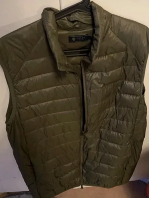 Olivgrön dunväst från Riley - Snygg olivgrön dunväst från Riley med quiltad design och hög krage. Västen har dragkedja framtill och är perfekt för lager-på-lager-stil. Tillverkad i lätt och värmande material som passar till många outfits.