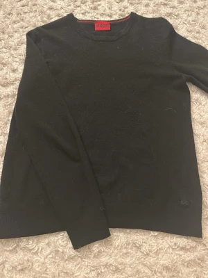 Svart stickad tröja från Hugo - Klassisk svart stickad tröja från Hugo med rund halsringning och diskret logga nertill. Tröjan har lång ärm och enkel, stilren design som passar till det mesta. Perfekt basplagg för garderoben. Håret som syns på tröjan är bara ull från mattan, tvättas såklart innan frakt.