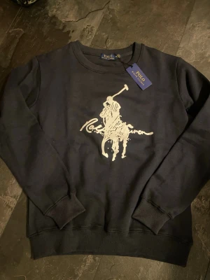 Marinblå sweatshirt från Polo Ralph Lauren M - Cool marinblå sweatshirt från Polo Ralph Lauren med stort vitt tryck av den klassiska polospelaren på bröstet. Tröjan har rund halsringning, ribbade muddar och är gjord i mjukt bomullsmaterial. Perfekt för en avslappnad och stilren look.