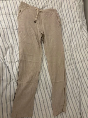 Beige linnebyxor xs - Snygga beige linnebyxor med avslappnad passform och elastisk midja med dragsko. Byxorna har sidfickor fram och två bakfickor med knapp. Perfekta för en chill och stilren look. Materialet är svalt och luftigt linne.