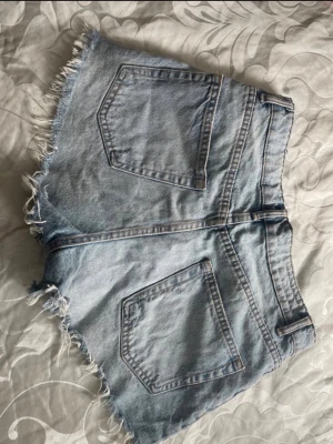 Ljusblåa högmidjade jeansshorts - Säljer ett par ljusblåa högmidjade jeansshorts i bomull med fransade kanter och klassiska bakfickor. Shortsen har normal passform och dragkedja framtill. Perfekta för sommaren och ger en avslappnad vibe.