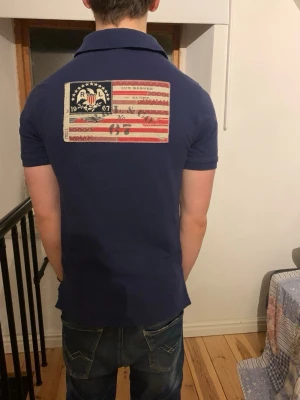 Vintage Marinblå pikétröja Polo Ralph Lauren - Säljer en marinblå pikétröja från Polo Ralph Lauren med krage och korta ärmar. På ryggen finns en stor patch med amerikansk flagga och textdetaljer. Klassisk och stilren modell som passar till mycket.