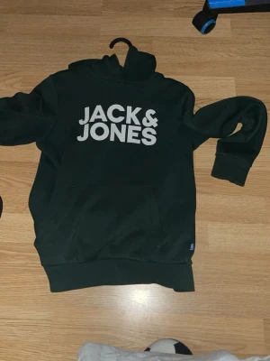 Mörkgrön hoodie från Jack & Jones - Snygg mörkgrön hoodie från Jack & Jones med stor vit logga på bröstet. Klassisk känguruficka framtill och huva. Tillverkad i mjukt bomullsmaterial som känns skönt mot huden. Perfekt för en avslappnad och trendig look.
